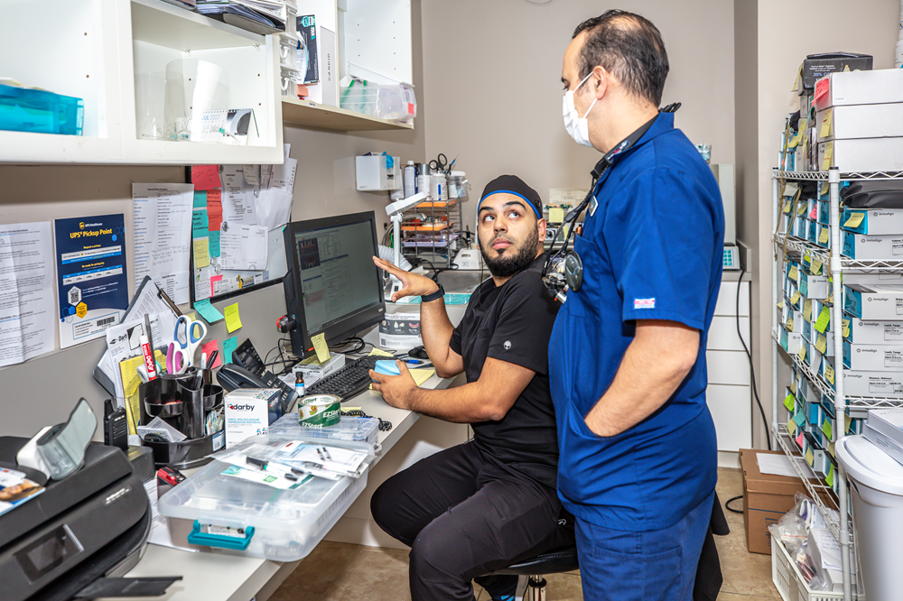 Two dental professionals in scrubs and masks discussing information on a computer in a well-stocked dental sterilization room, surrounded by organized supplies and equipment - Dental Assisting Institute