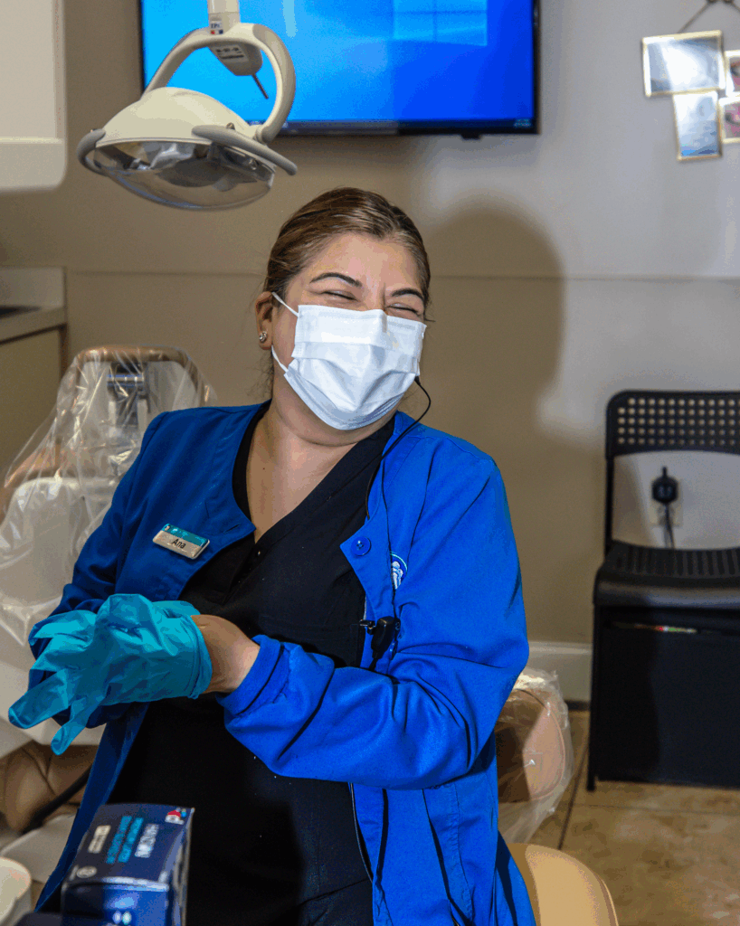 Female in a blue jacket, face mask, and gloves smiling while preparing for a procedure in a clean dental operatory with overhead lighting and a TV screen - Dental Assisting Institute